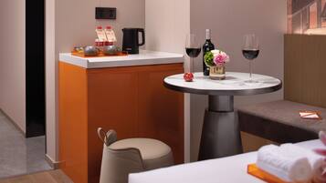 A modern hotel room with a round table, a minibar, a sofa, and a small round table with a bottle of wine and a glass.