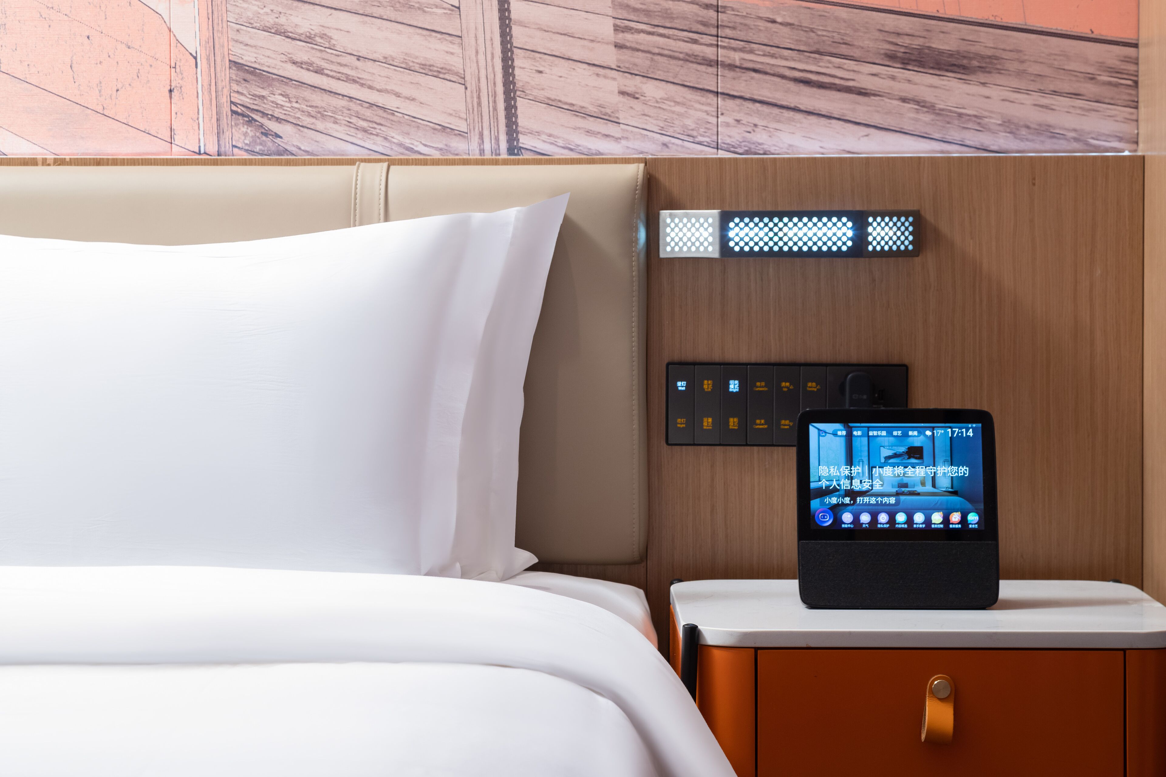 Ein Hotelzimmer mit einem Bett, einem Nachttisch mit einem digitalen Gerät, das Zimmerinformationen anzeigt, und einem Holz-Kopfteil.