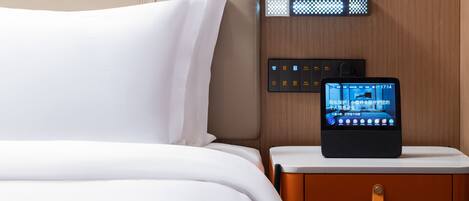 Ein Hotelzimmer mit einem Bett, einem Nachttisch mit einem digitalen Gerät, das Zimmerinformationen anzeigt, und einem Holz-Kopfteil.