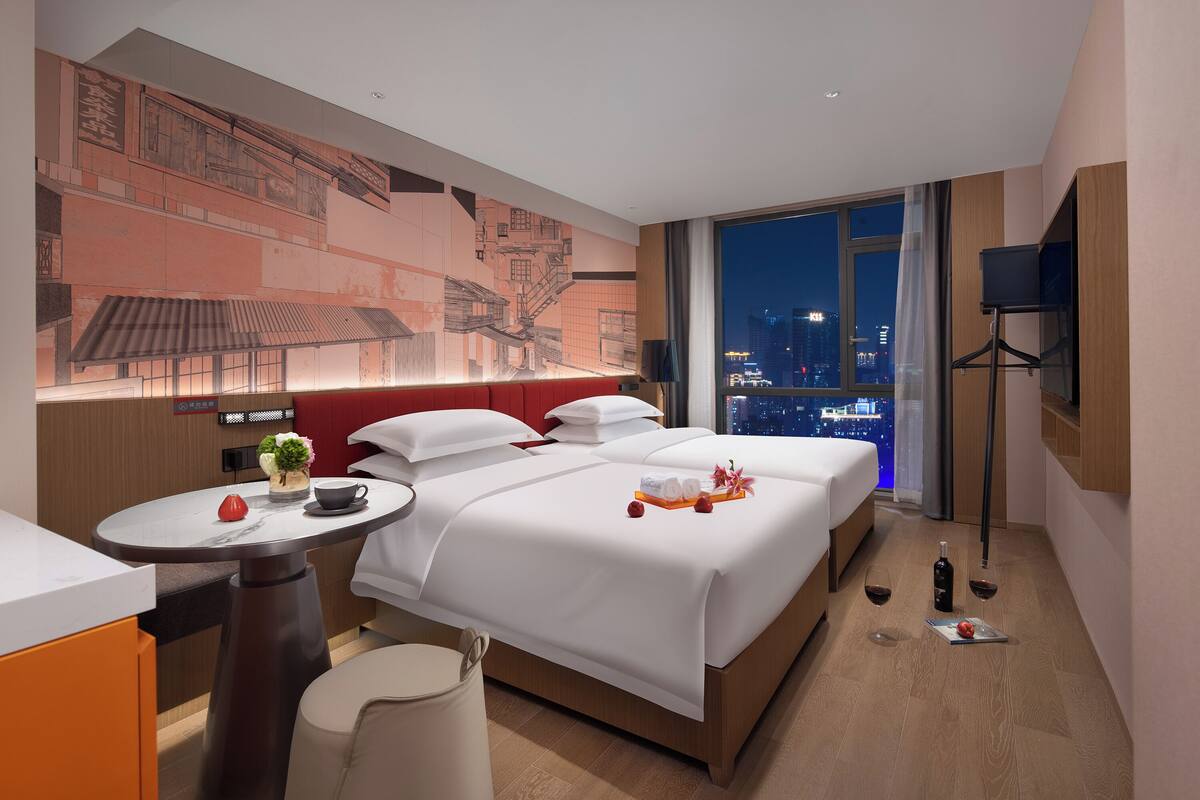 Habitación de hotel con una cama grande, una mesita con un florero y una taza, un televisor sobre un mueble bajo, y vistas a la ciudad a través de la ventana.