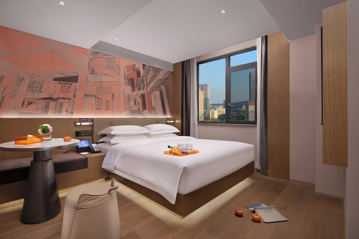 Una habitación de hotel moderna con una cama grande, un escritorio con computadora, una mesita con planta y un amplio ventanal con vista a la ciudad.