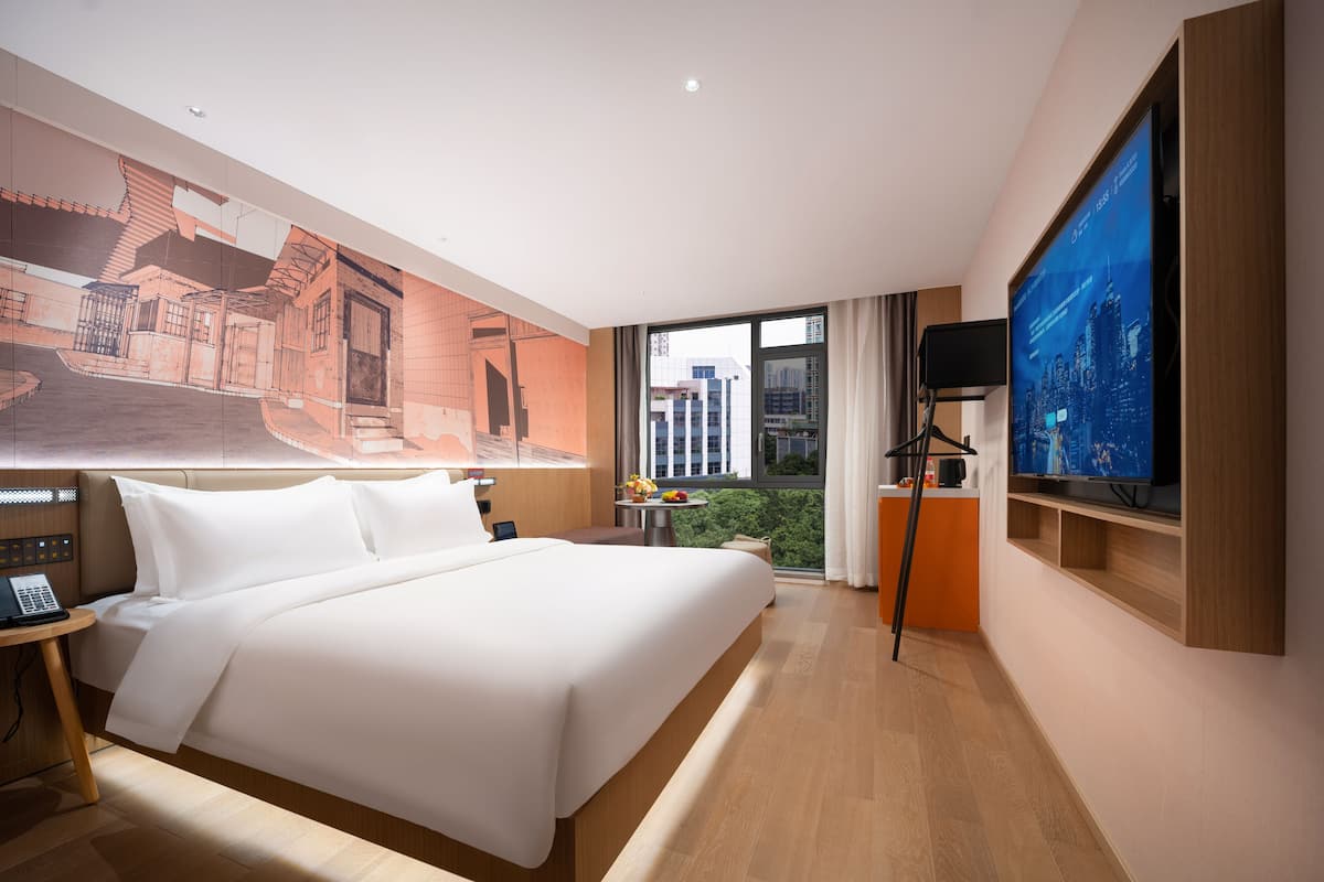 Habitación de hotel con una cama grande, un escritorio con una computadora portátil, un televisor y vista a la ciudad por la ventana.