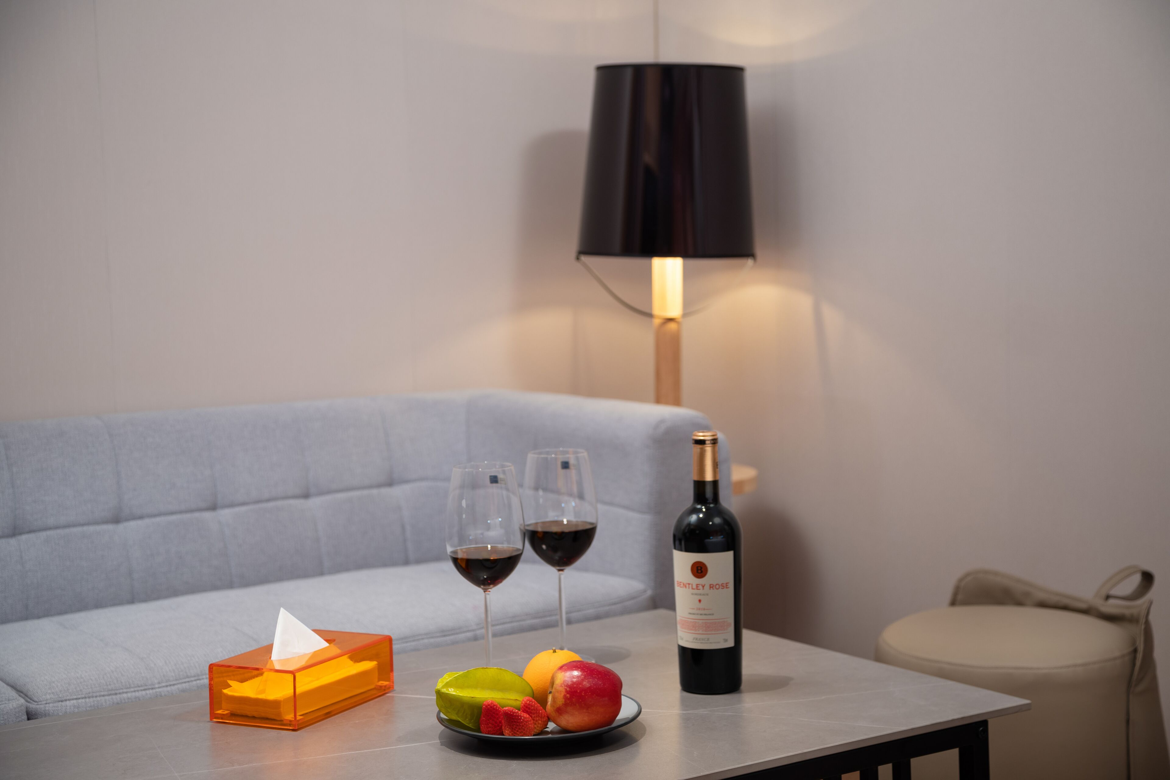 A table with a bottle of wine, two glasses of red wine, a plate with fruit, and a tissue box.