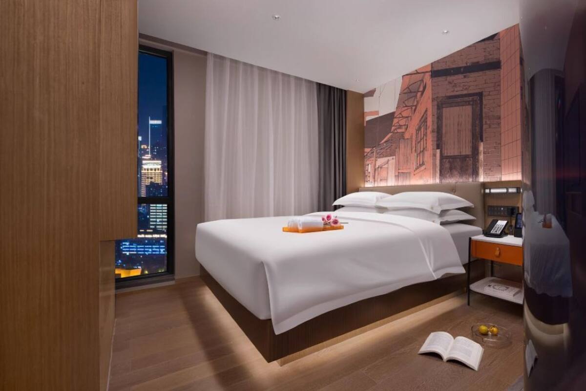 Habitación de hotel moderna con una cama grande, una mesita de noche, vistas a la ciudad y un ventanal con cortinas.