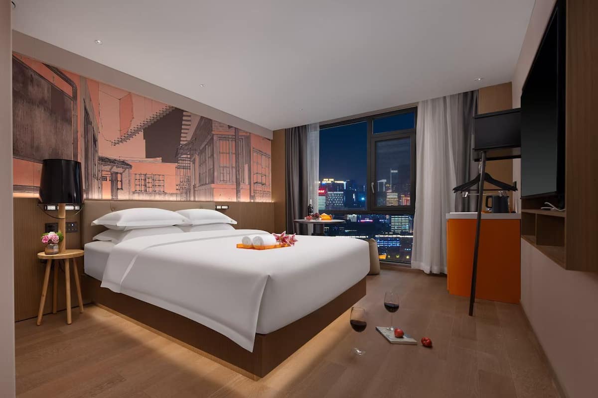 Una habitación de hotel con una cama grande, vistas a la ciudad, un escritorio con televisor y una mesita de noche con lámpara.