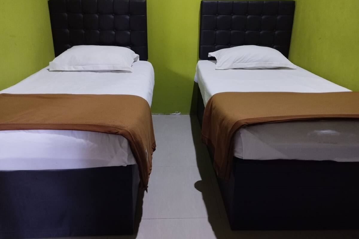 Deux lits simples, dotés de têtes de lit noires et de draps blancs, sont placés contre un mur vert, dans une chambre au décor simple.
