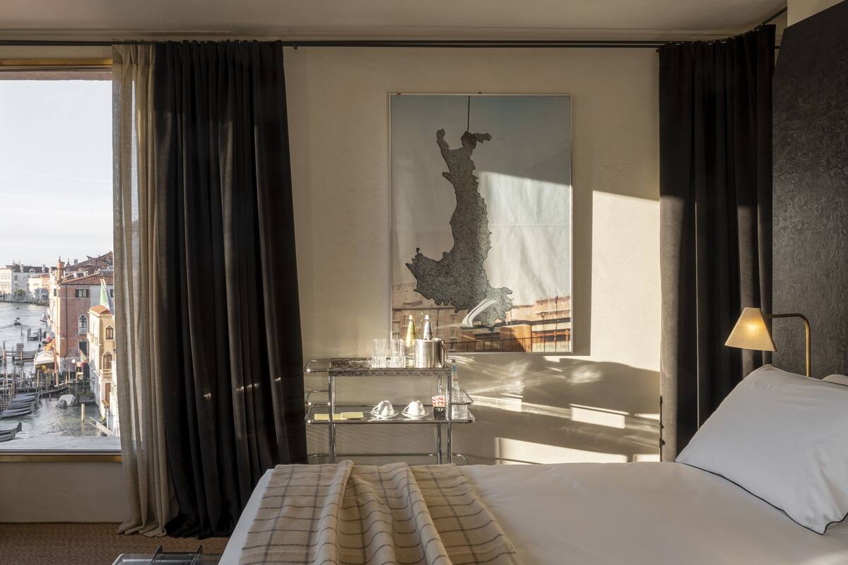 Een hotelkamer met een bed, een bureau met lamp, een groot raam met gordijnen en uitzicht op de stad.