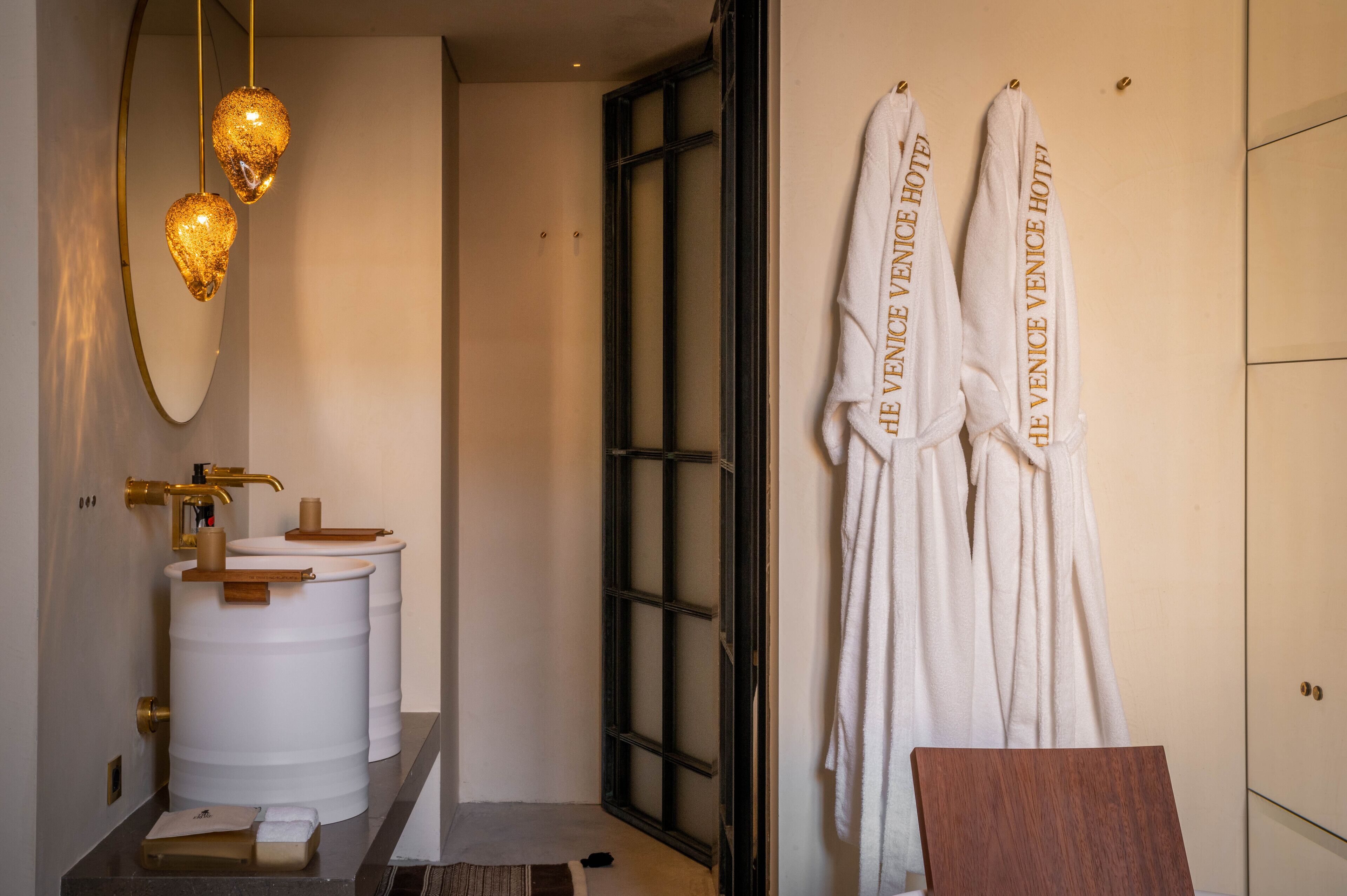 Ein Badezimmer mit einem weißen Waschbecken, zwei goldenen Pendelleuchten und zwei weißen Handtüchern, die an der Wand aufgehängt sind. Auf den Handtüchern steht 'HE VENICE HOTEL'.