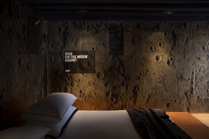 Ein Zimmer mit einer strukturierten Wand, die die Mondoberfläche ähnelt, einem Bett mit Kissen und einem Schild mit der Aufschrift 'MAN ON THE MOON AGAIN?'