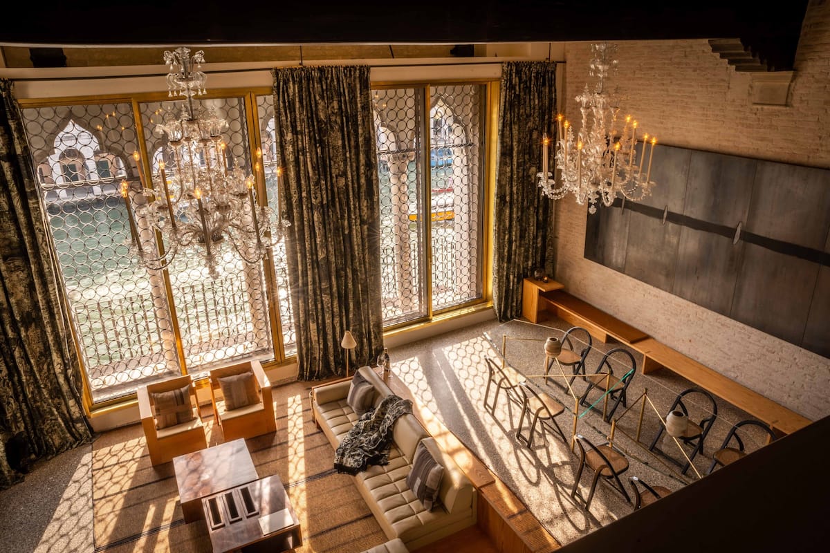 Een ruime kamer met een groot raam, een bank, een bijzettafeltje en een kroonluchter.