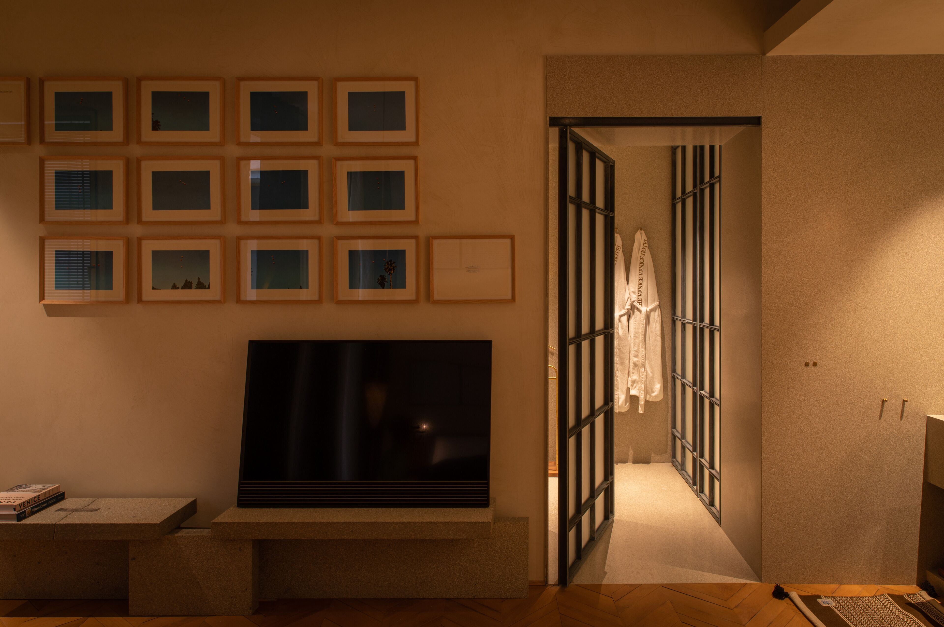 A hotel room with a large flat-screen TV mounted on the wall, a series of framed pictures above it, and a sliding door leading to a bathroom.