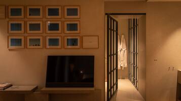 A hotel room with a large flat-screen TV mounted on the wall, a series of framed pictures above it, and a sliding door leading to a bathroom.