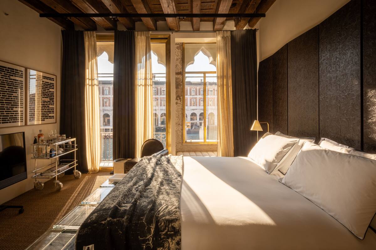 Een hotelkamer met een bed, een bureau met lamp, een raam met gordijnen en uitzicht op een gebouw.