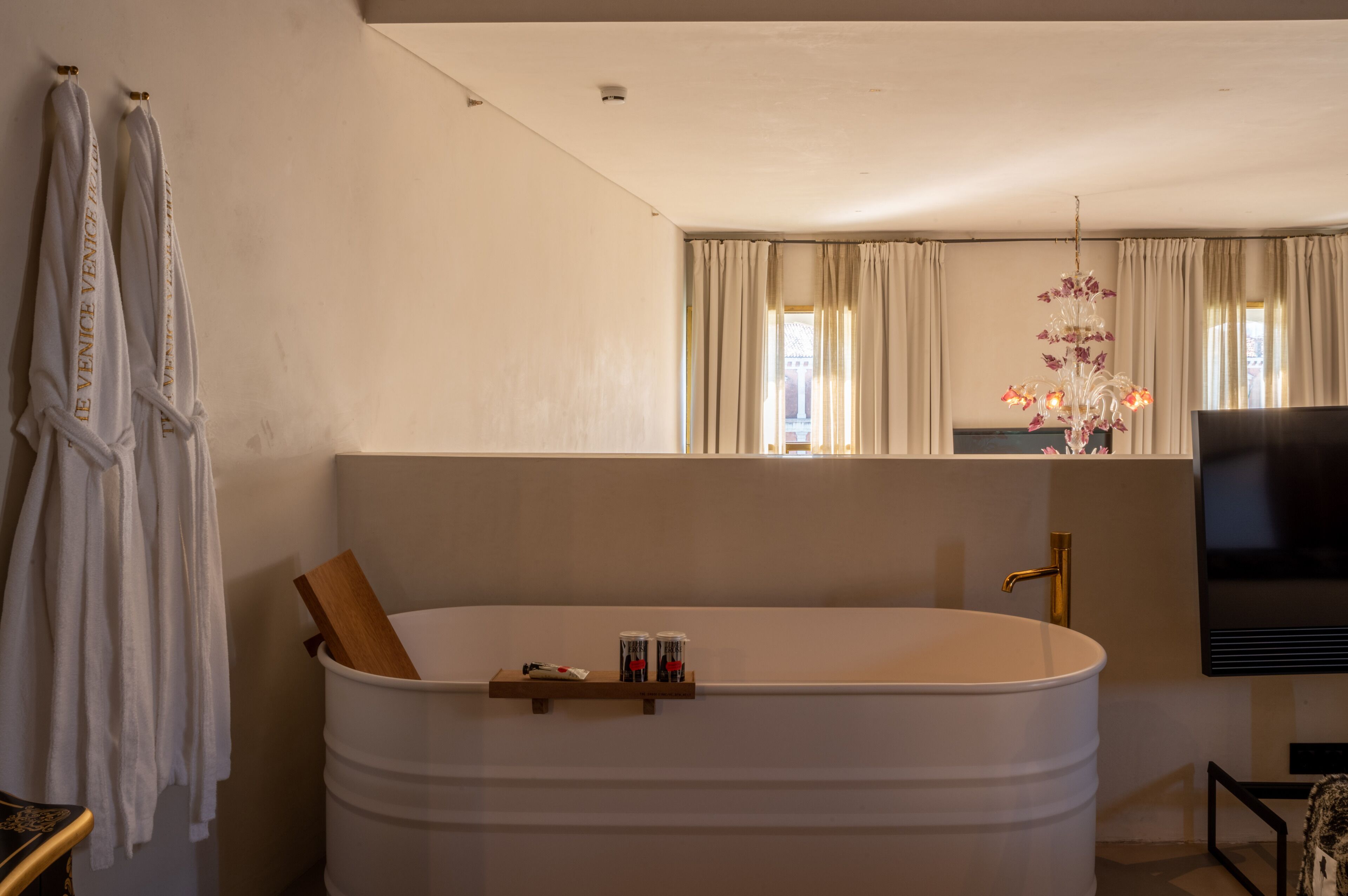 Un baño moderno con una bañera grande, un estante de madera con artículos de aseo, un televisor y un espejo que refleja la habitación.