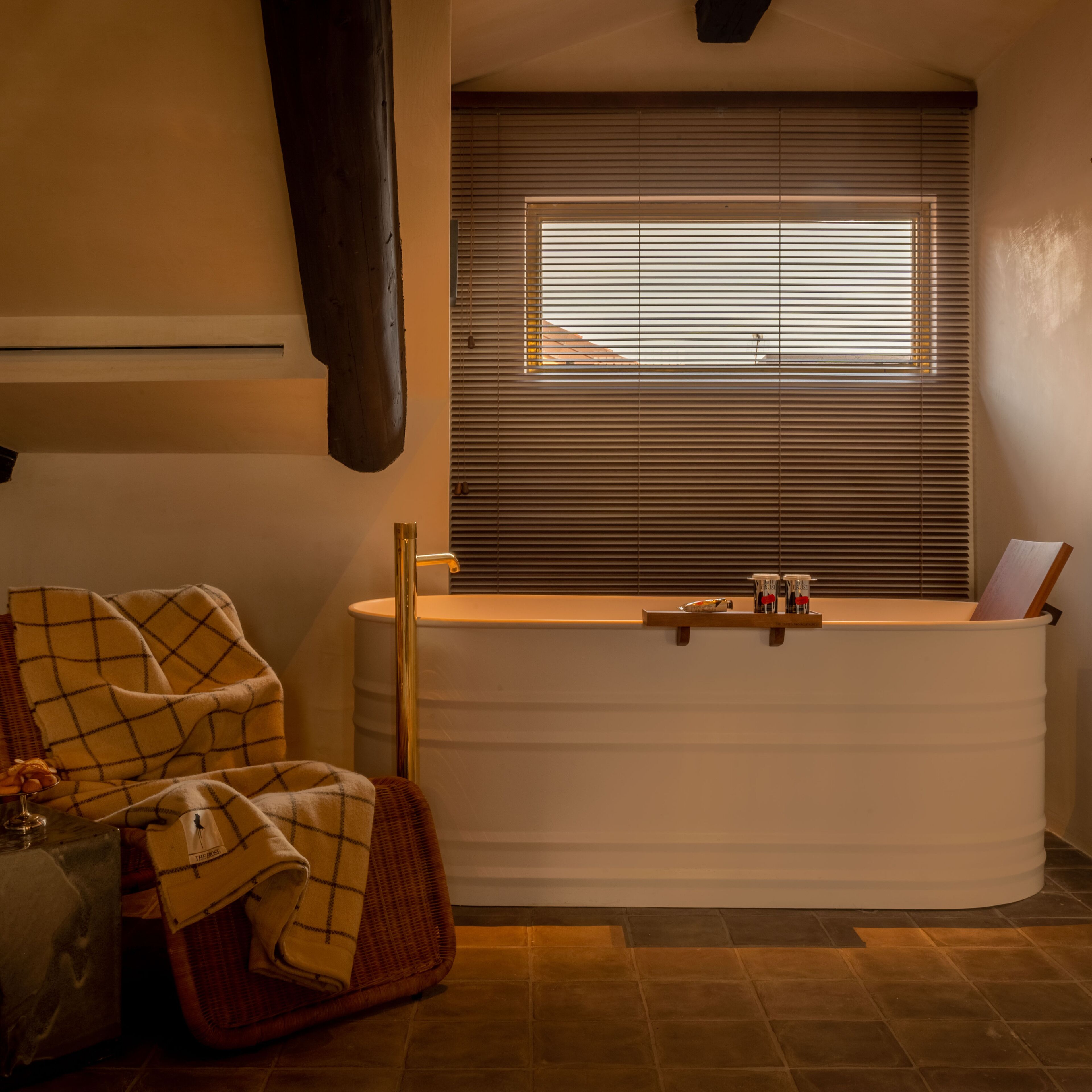 Une salle de bain avec une baignoire, une fenêtre avec des stores, un porte-serviettes et une étagère avec des articles de toilette.
