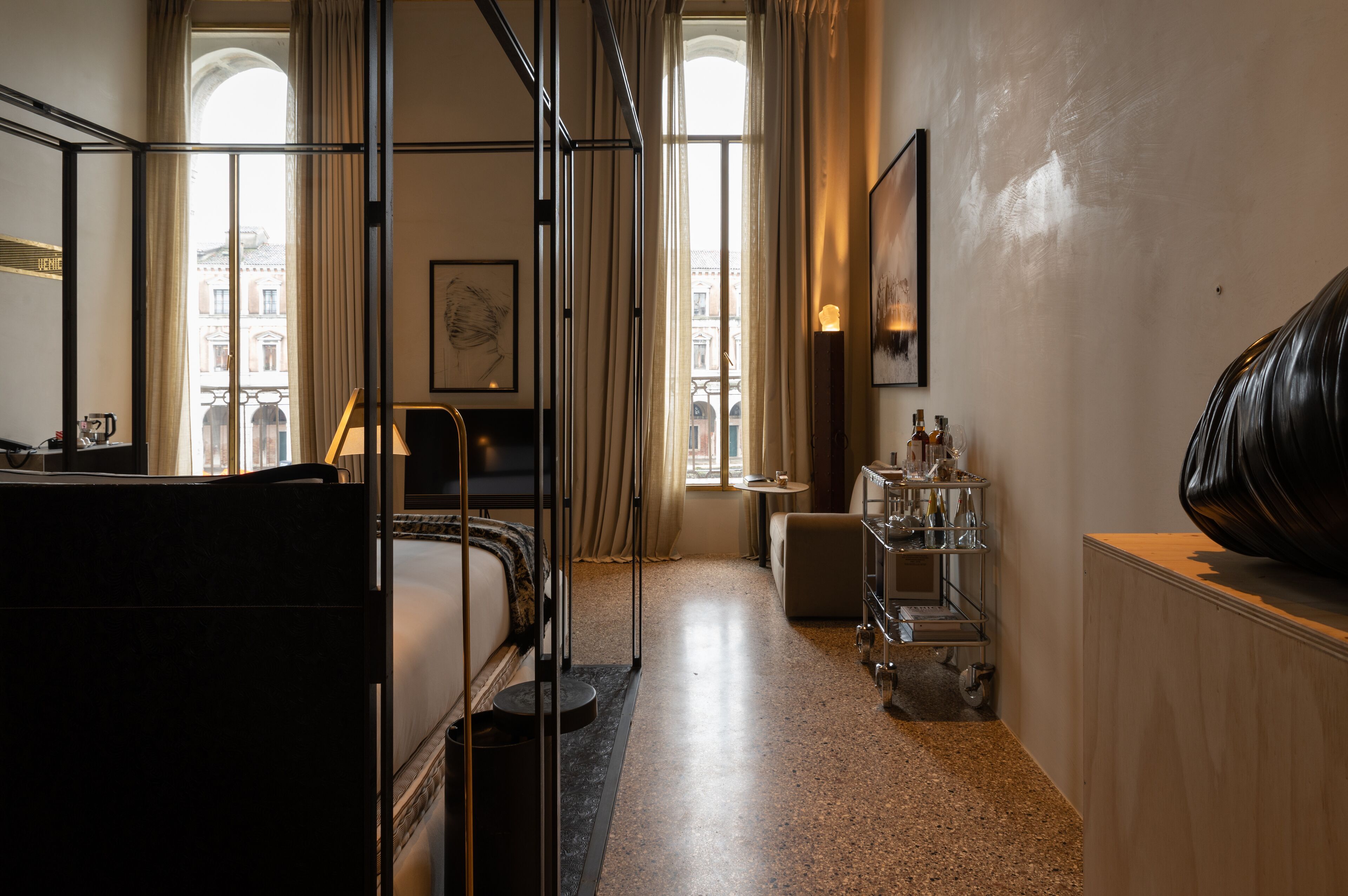 Une chambre d’hôtel avec un lit, un bureau, une chaise, une petite table, une étagère avec des bouteilles et une grande fenêtre avec des rideaux.