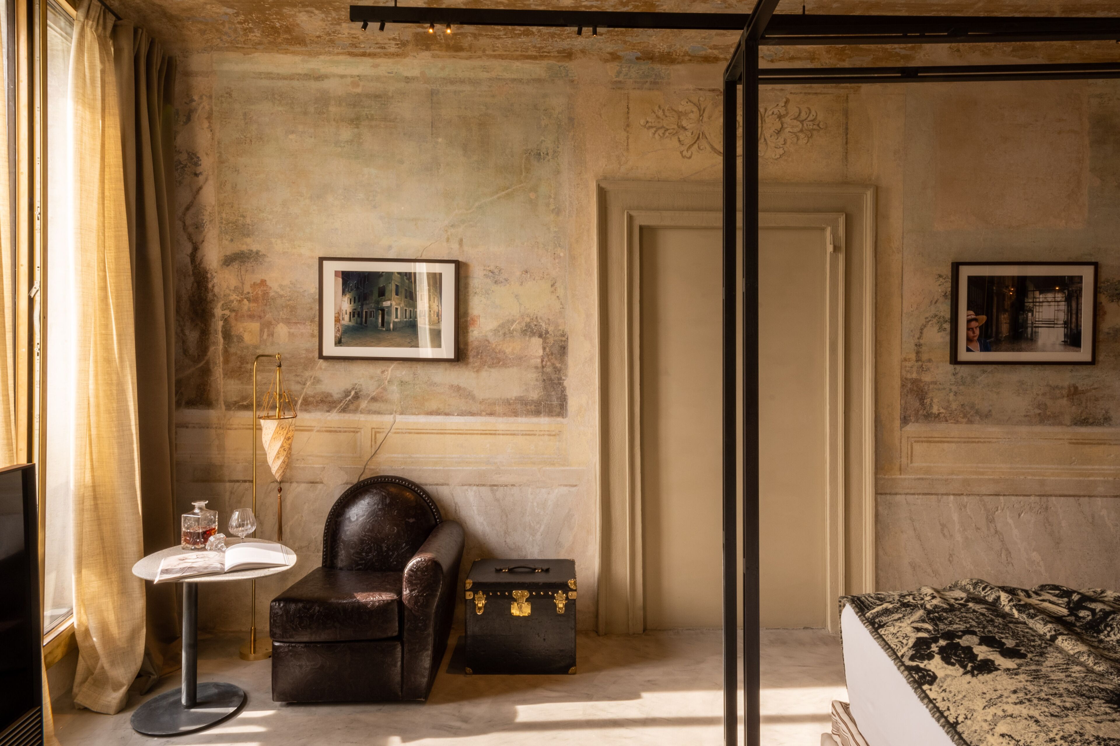 A room with a leather armchair, a small round table with a book and glasses, a vintage suitcase, and two framed pictures on the wall.