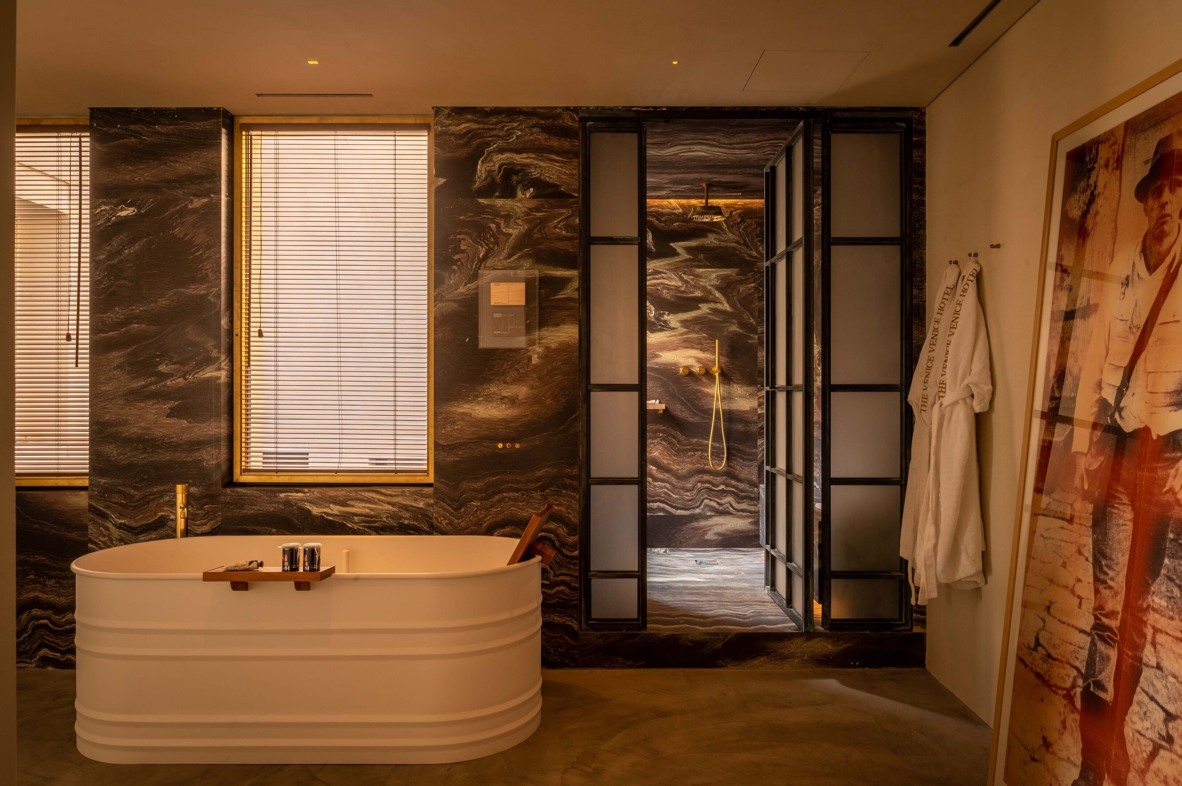 Un baño moderno con una bañera independiente, paredes de mármol y un amplio ventanal con persianas.