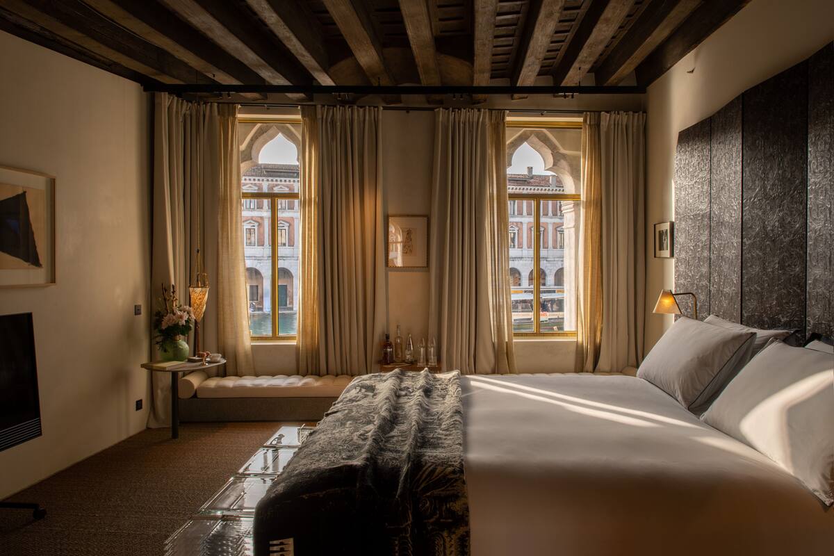 Hotelkamer met een groot bed, twee ramen met gordijnen, een kleine tafel met een vaas en een tv aan de muur.