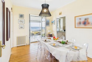 A dining area with a table set for four, clear chairs, a white tablecloth, and a view of the sea through an open door.