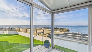 Uitzicht vanaf een balkon met een glazen deur, uitkijkend op een strand met een rotsachtige kustlijn en een heldere lucht.