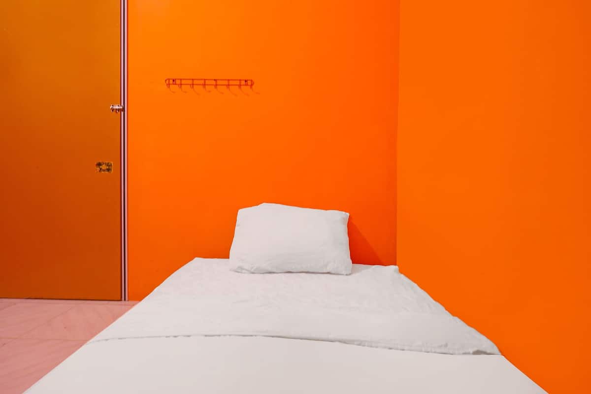 A room with a bed against an orange wall, a door with a handle, and a small shelf with a hook.