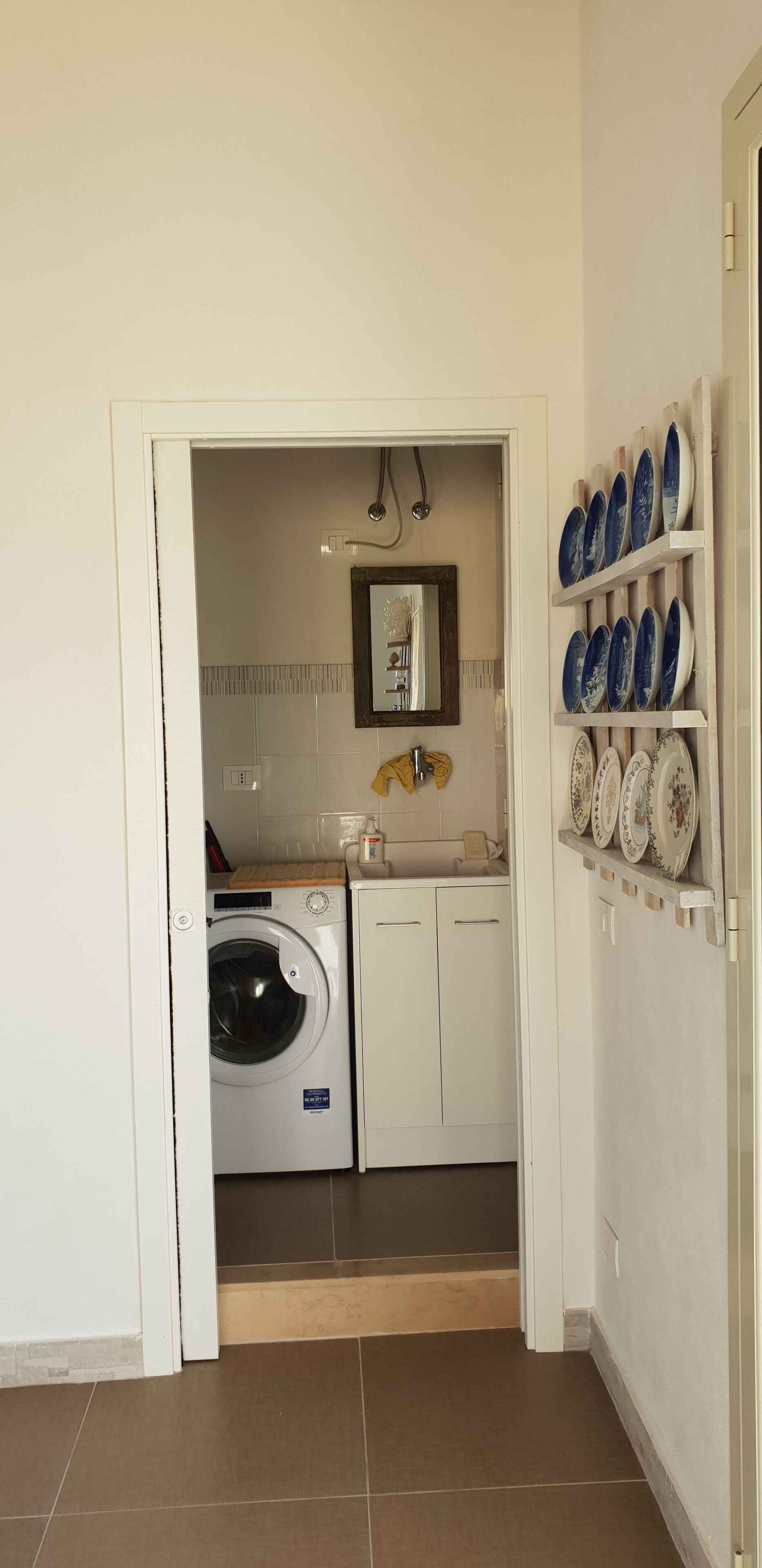 A compact kitchen with a washing machine, a sink, and a shelf with plates.