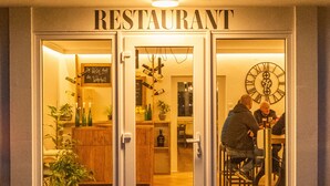 A restaurant with a sign above the entrance, a clock on the wall, and a person sitting inside. - Hotel Fuchs und Hase (Rosegg)