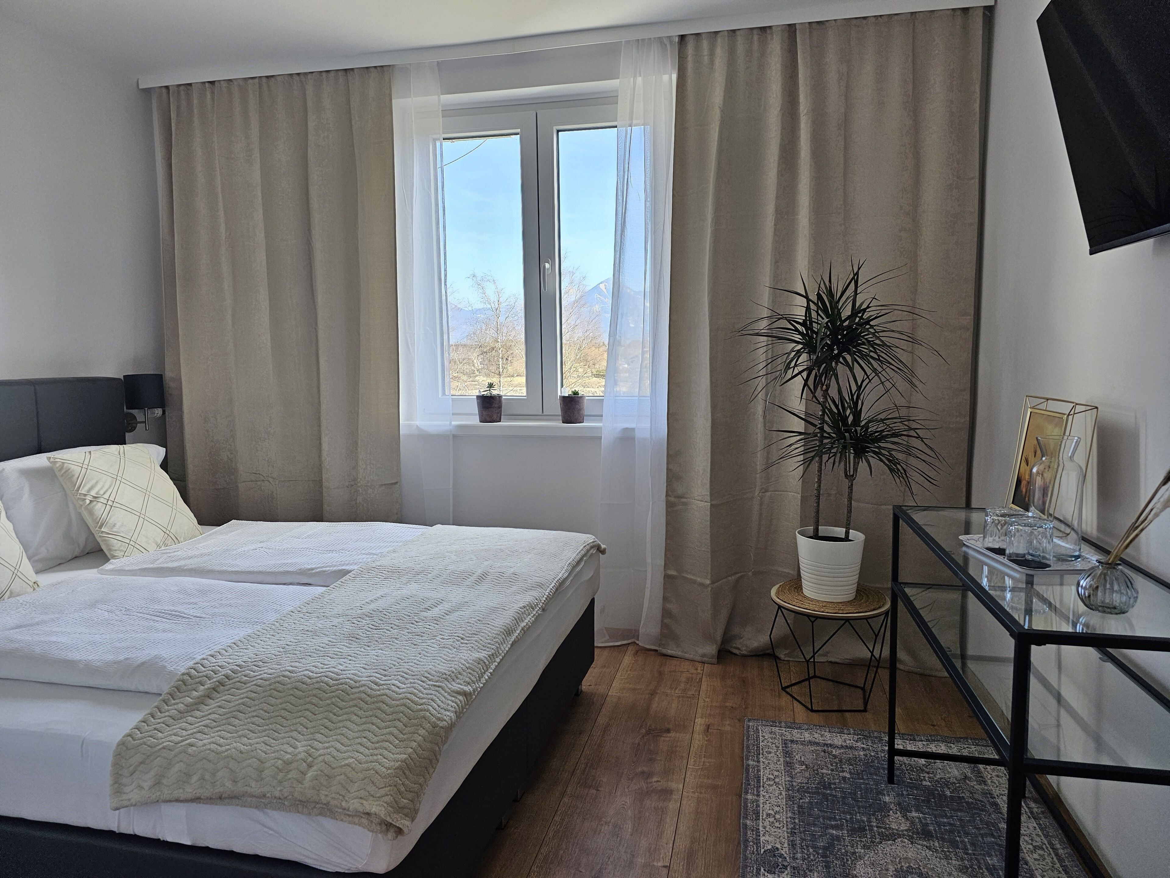 Een slaapkamer met een bed, een raam met gordijnen, een potplant, een glazen tafel met een vaas en een lijst met een foto.