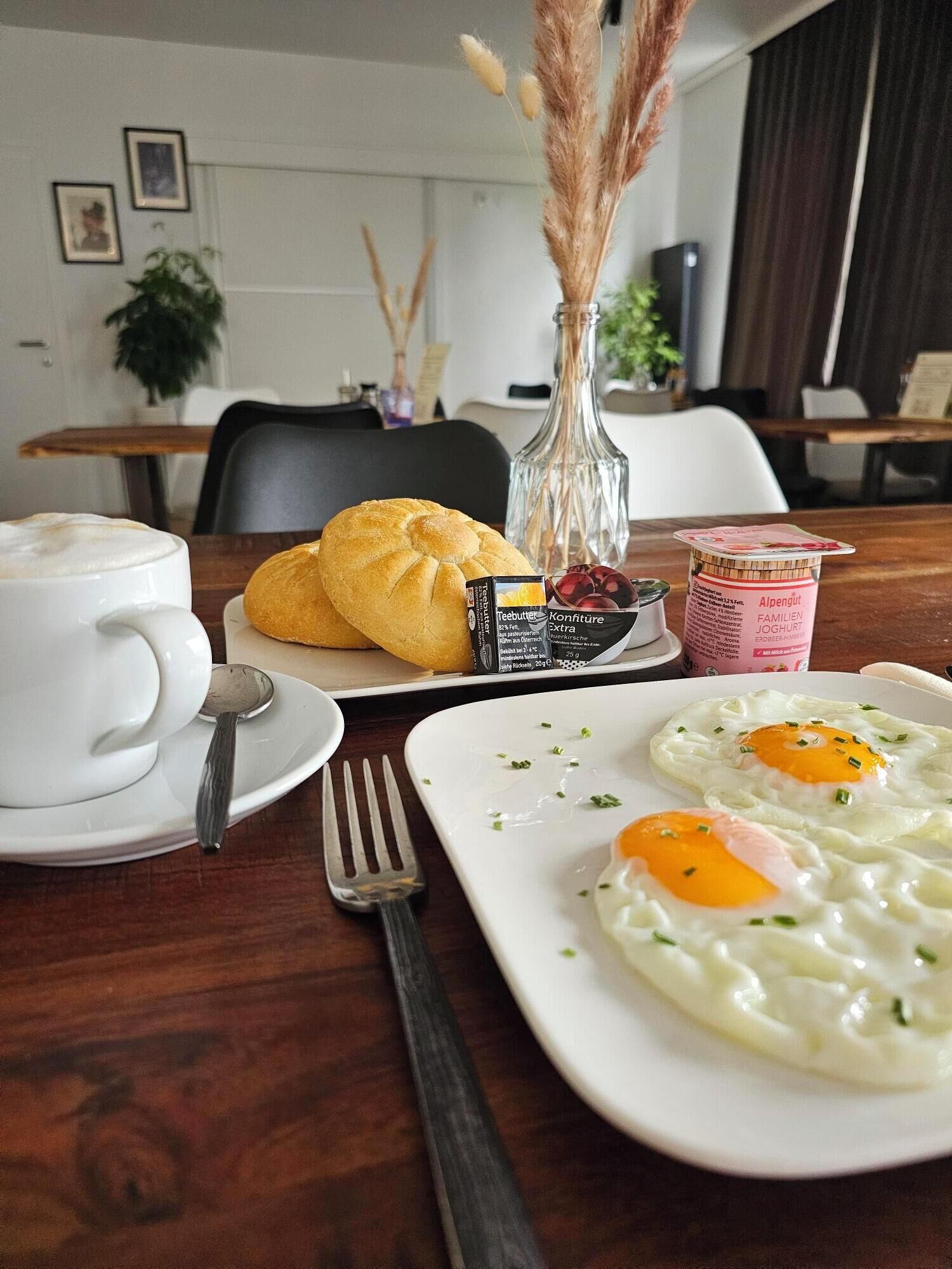 A breakfast table with a croissant, a glass of coffee, a plate of fried eggs, and a container of yogurt.