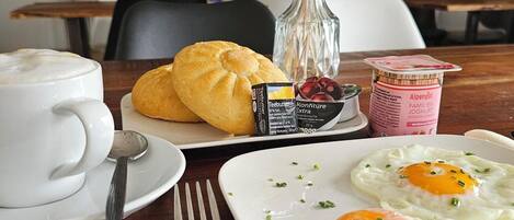 A breakfast table with a croissant, a glass of coffee, a plate of fried eggs, and a container of yogurt.