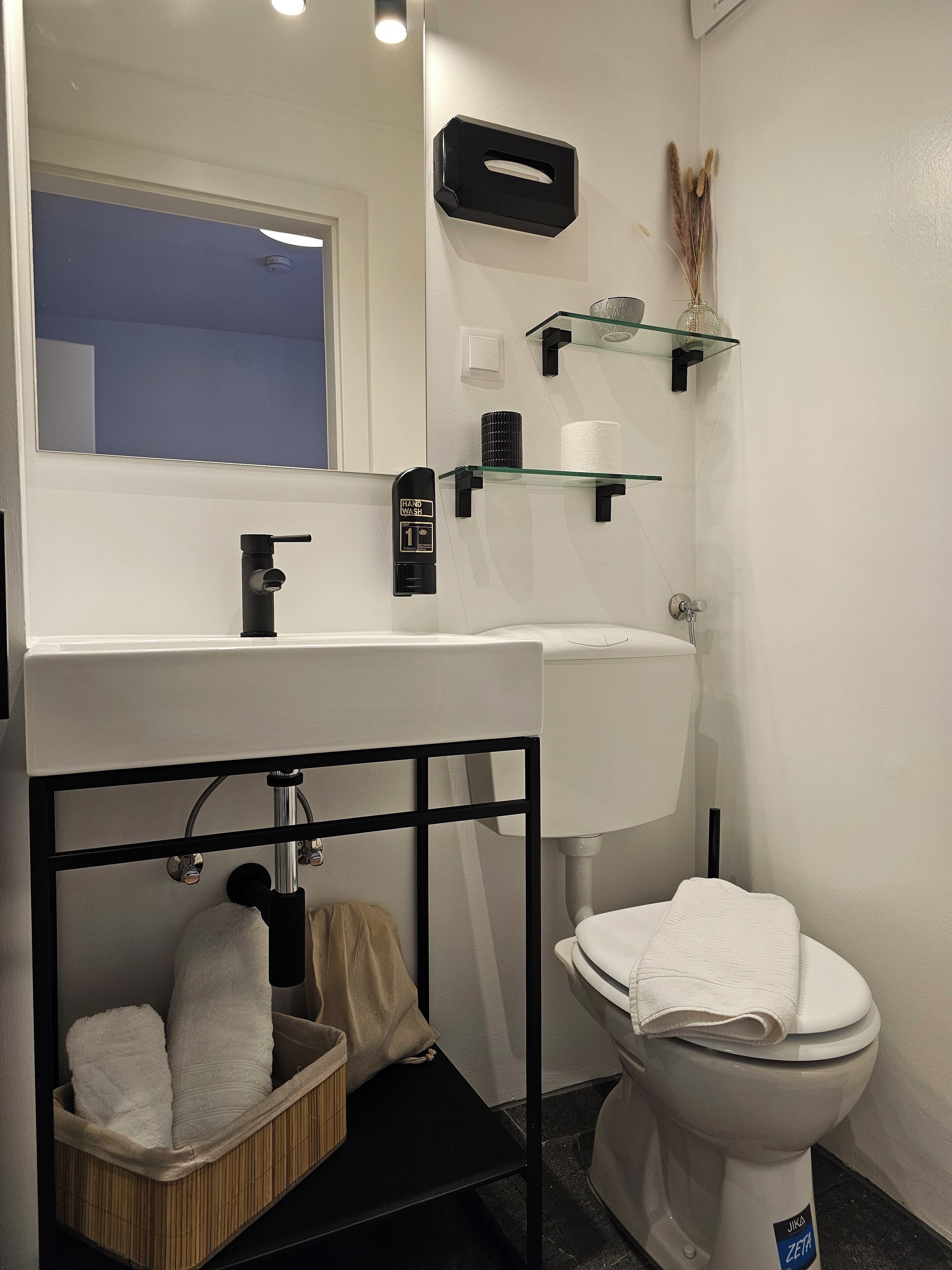 A modern bathroom with a white sink, black faucet, and toilet. There are glass shelves above the sink and a black metal frame below the sink.