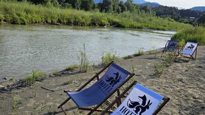 Three beach chairs with "HOTEL" logos on them, placed on a sandy beach beside a river with mountains in the background. - Hotel Fuchs und Hase (Rosegg)