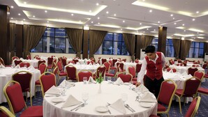 A banquet hall with round tables, red chairs, and a waiter attending to a table.