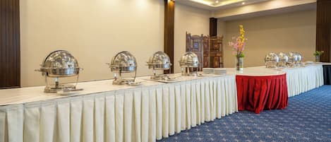 A banquet hall with a long table covered with a white tablecloth and a red table runner, surrounded by metal food service equipment.