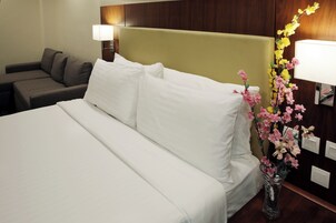 Una habitación de hotel con cama, sofá, mesita de noche con lámpara y un florero con flores.