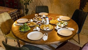 Une table de salle à manger ronde en bois, dressée avec différents plats, assiettes et couverts, entourée de chaises dans une pièce aux murs texturés et agrémentée d’une plante.