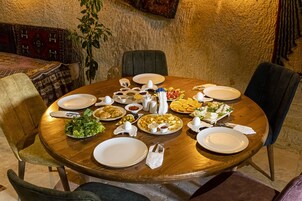 A round wooden dining table set with various dishes, plates, and utensils, surrounded by chairs in a room with textured walls and a plant.