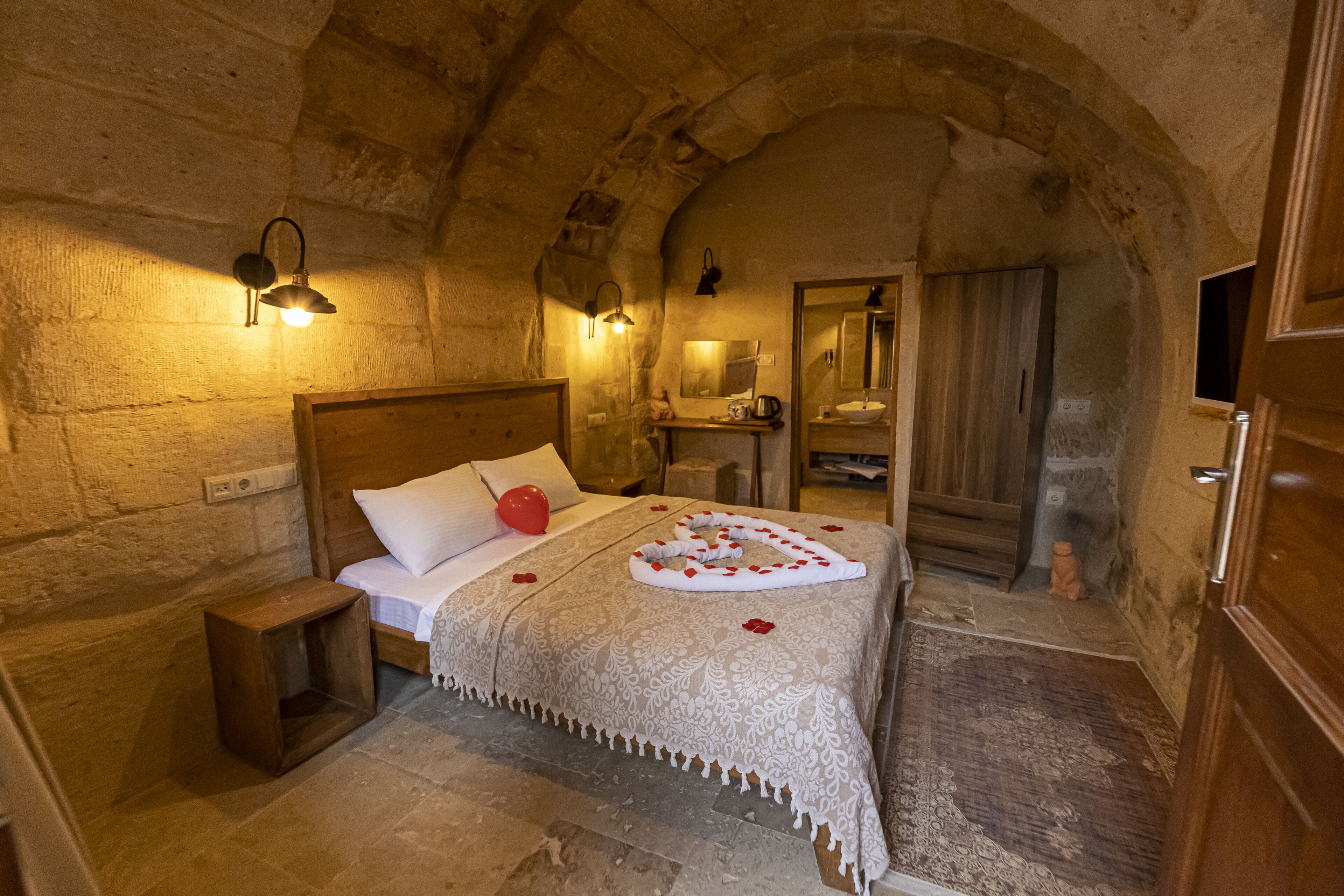 A stone-walled bedroom with a wooden bed, a nightstand, a small stool, and a bathroom visible through an arched doorway.