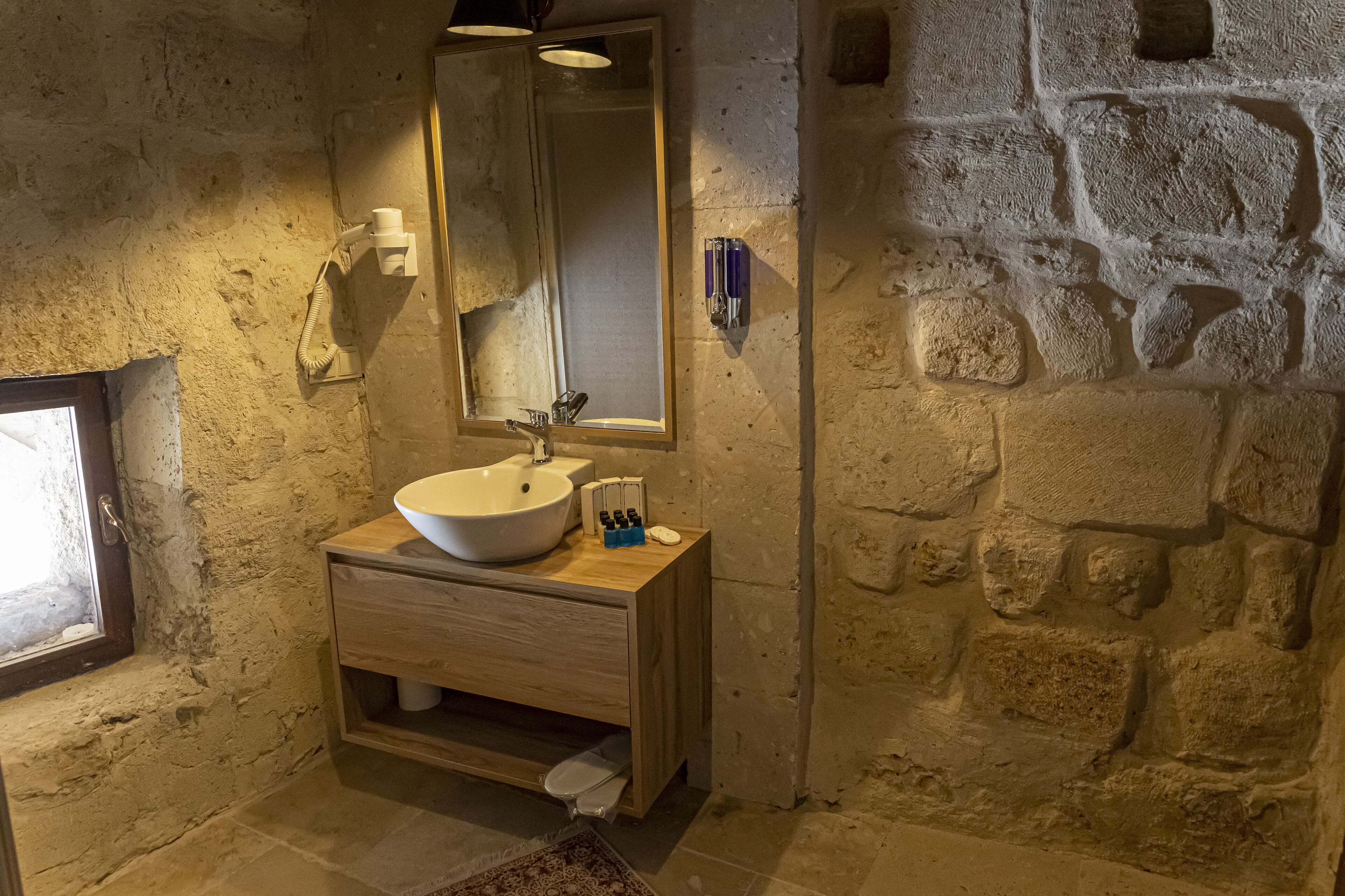 A bathroom with a stone wall, a wooden vanity, a white sink, a mirror, and a window.