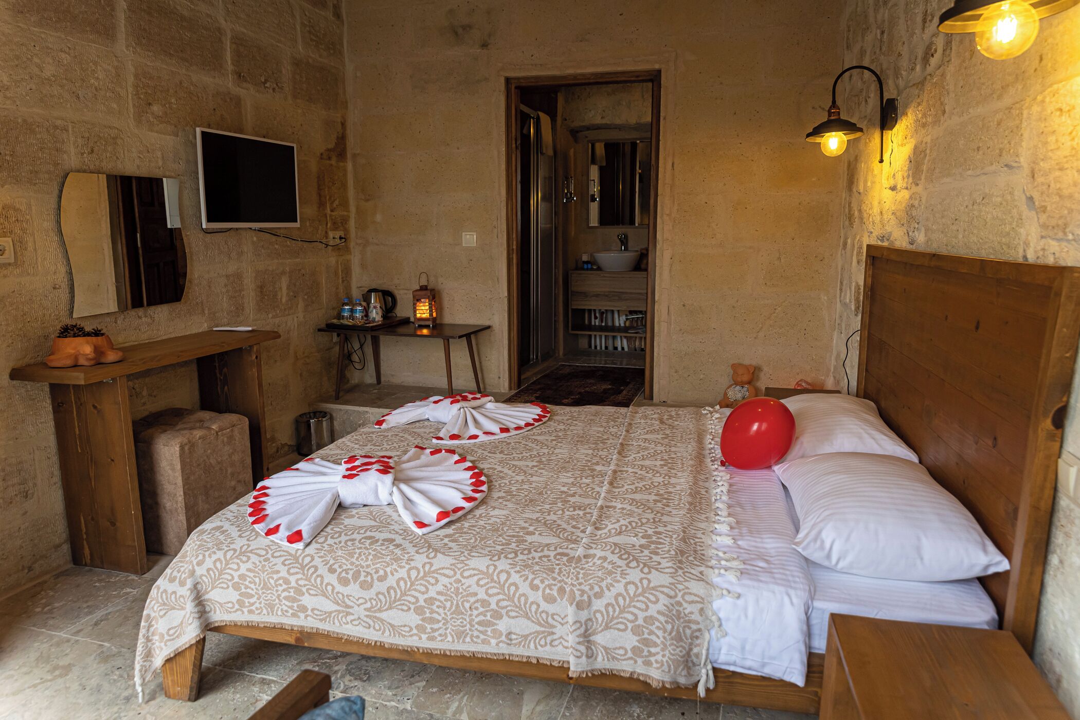 A room with a bed, a wooden headboard, a bedside table, a small table with a lantern, a mirror, and a television mounted on the wall.