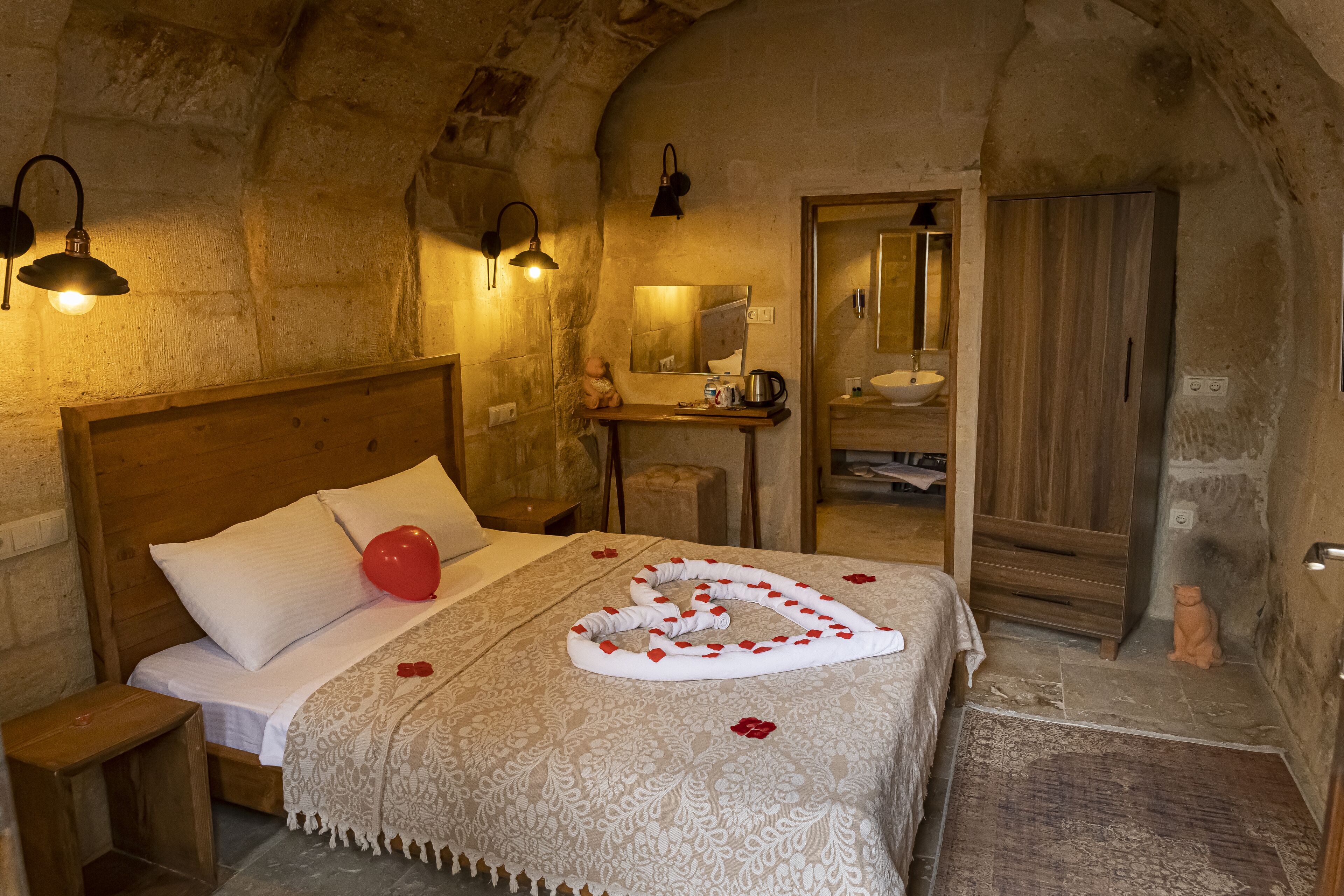 Un dormitorio acogedor con cabecera de madera, cama con colcha blanca y un globo rojo, mesita de noche de madera y pared de piedra.