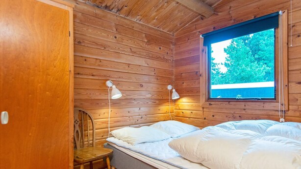 A wooden cabin room with a bed, two wall-mounted lamps, a window with a view of greenery, and a door. - 6 Person Holiday Home in Henne (Henne)