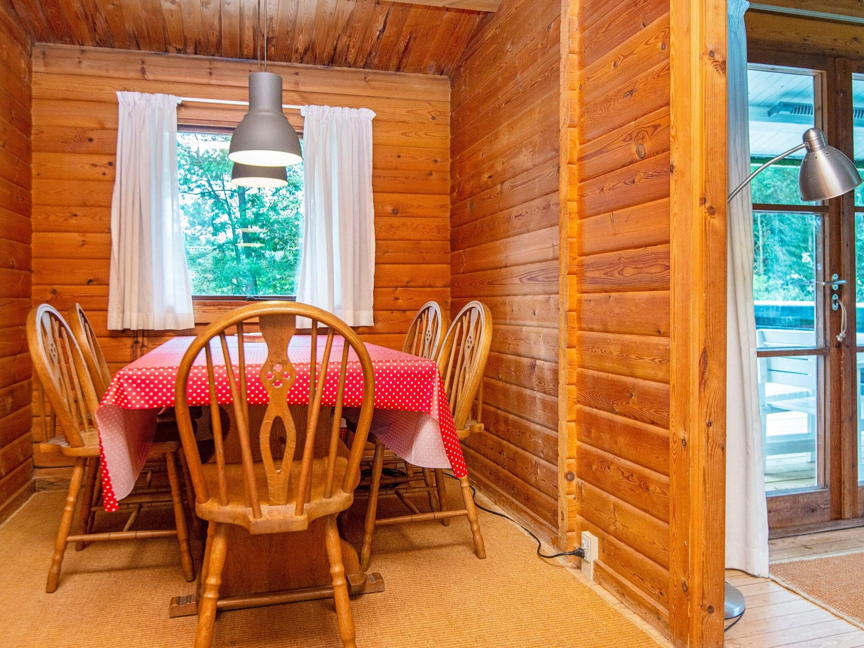 Un interno in legno con un tavolo da pranzo imbandito per quattro persone, sedie in legno, una lampada a sospensione e una finestra con tende.