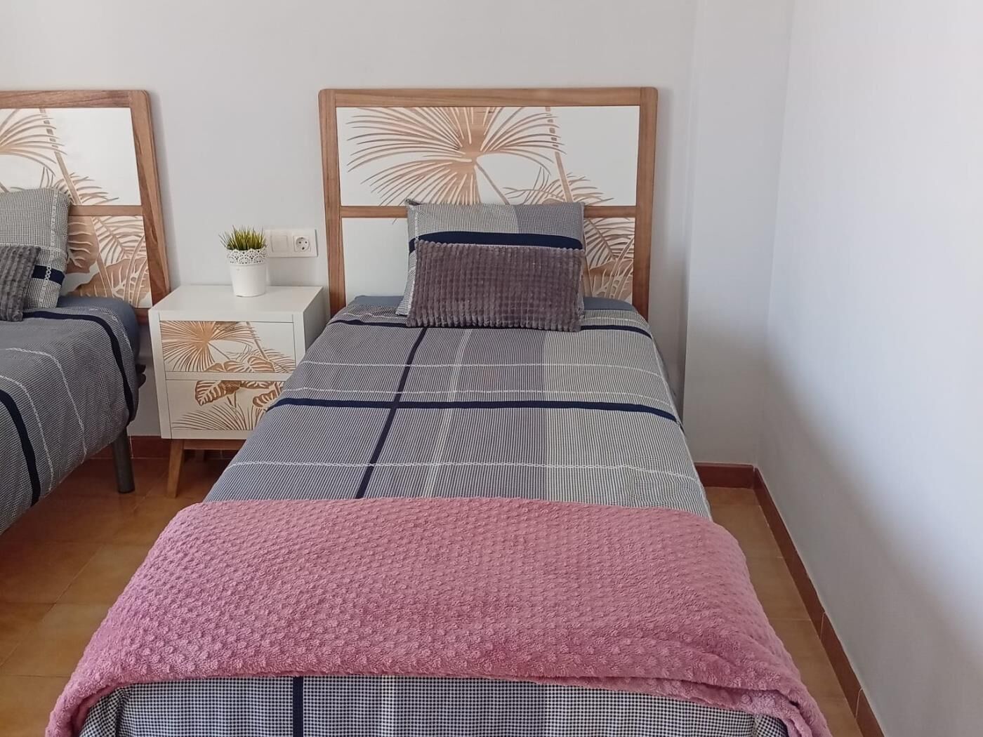 A double bed with a wooden headboard, a bedside table with a plant, and a patterned wall.