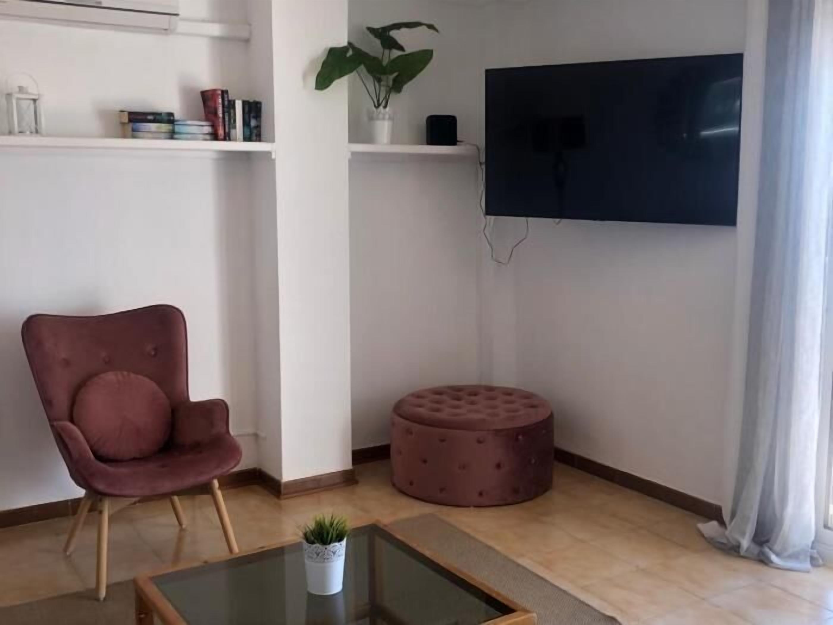 A living room with a red chair, a glass coffee table, a round ottoman, a potted plant, and a flat-screen TV mounted on the wall.