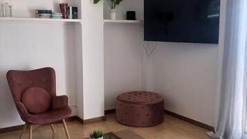 A living room with a red chair, a glass coffee table, a round ottoman, a potted plant, and a flat-screen TV mounted on the wall.