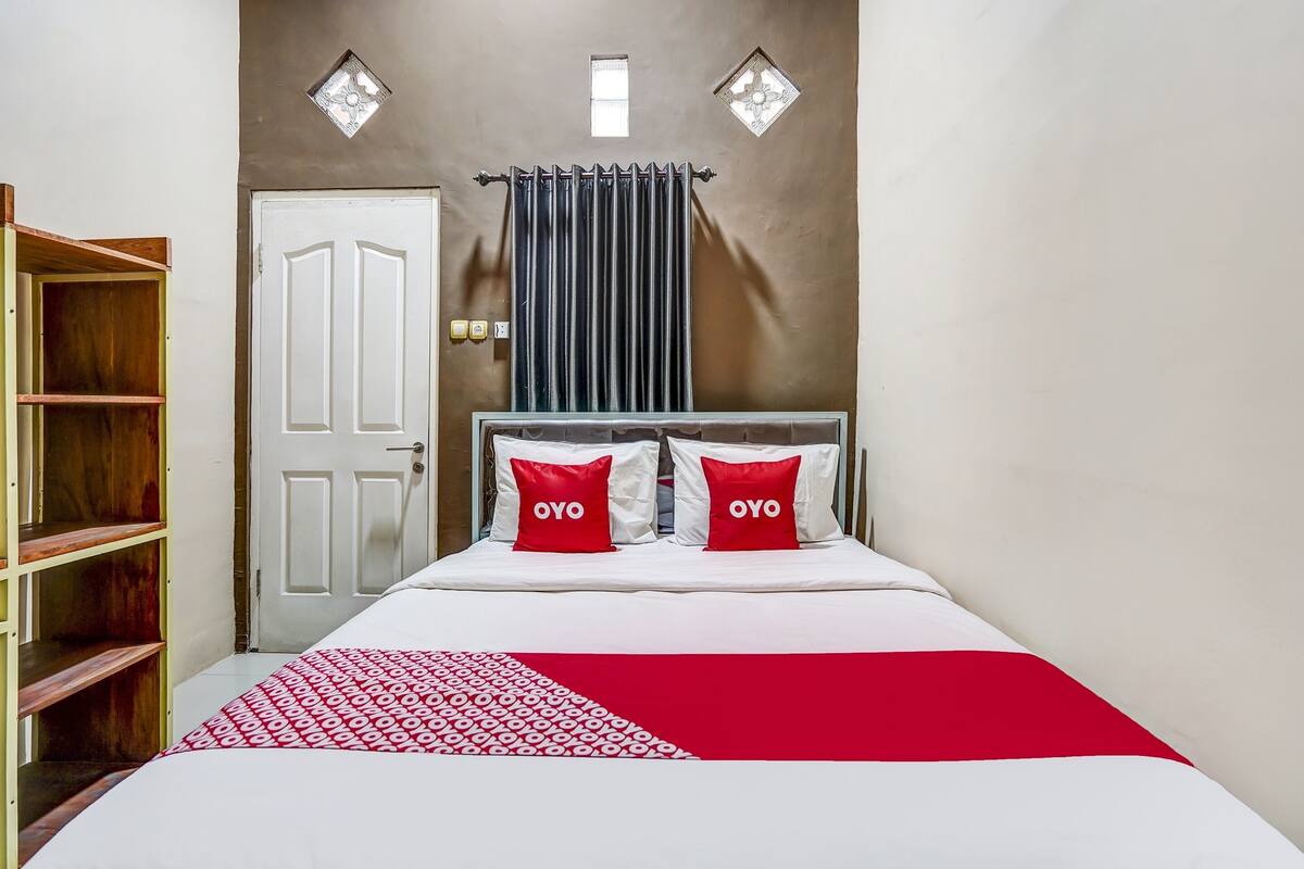 A bedroom with a bed, two pillows with the word "OYO" on them, a red and white patterned bedspread, a white door, and a wooden wardrobe.