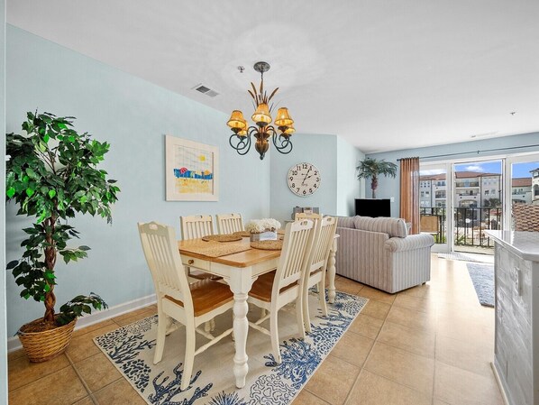A dining area with a wooden table and chairs, a chandelier, a wall clock, and a large window with a view of buildings.