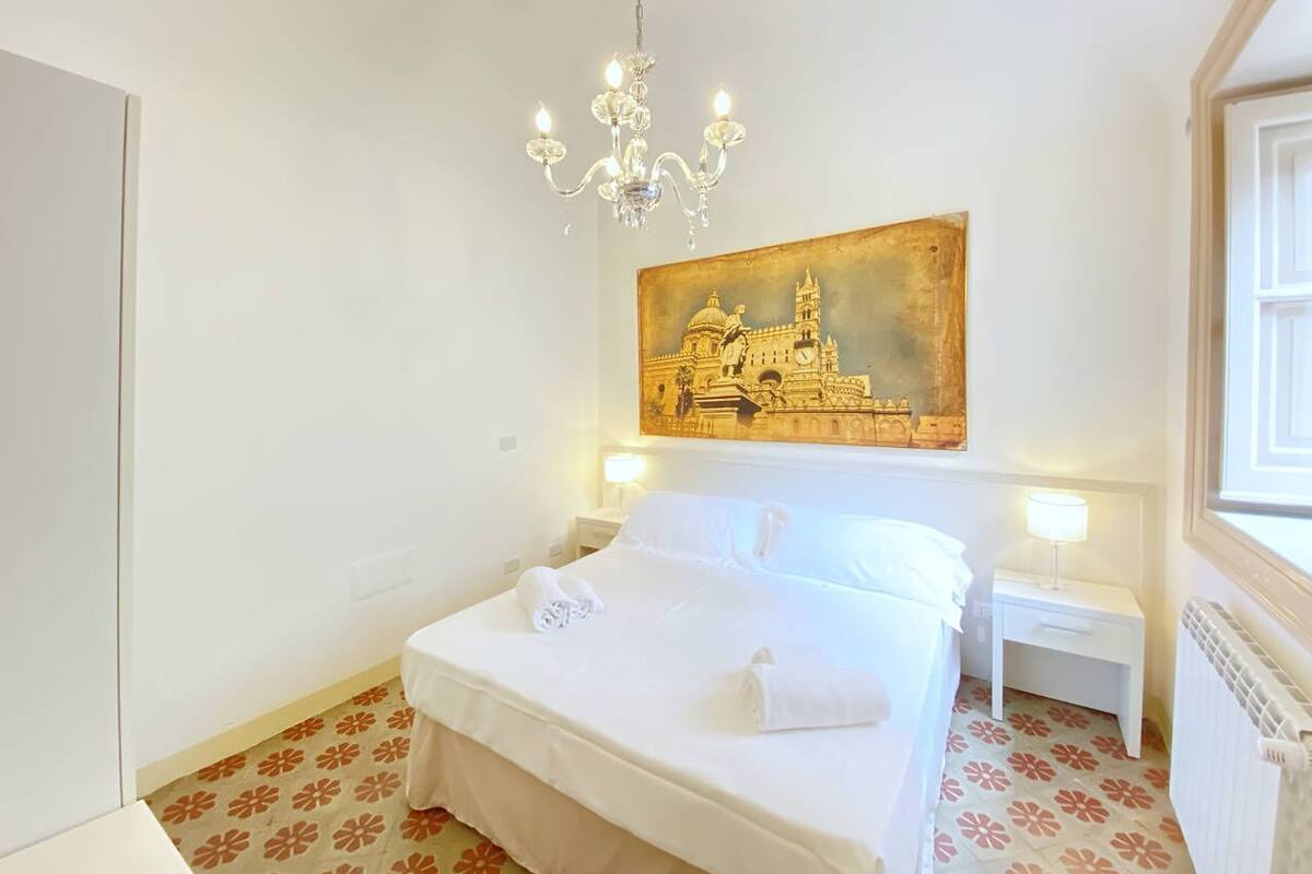 A neatly made bed with white linens and a rolled towel, a chandelier, a nightstand with a lamp, and a painting of a building on the wall.